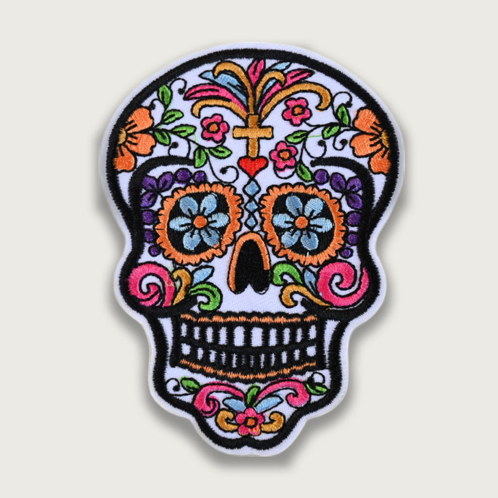 Colorful embroidered sugar skull patch inspired by Día de los Muertos, featuring floral patterns, a cross, and vibrant decorative details